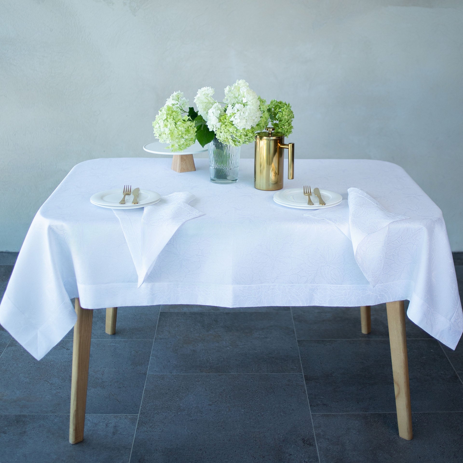 Dining table set with white tablecloth, plates, silverware, and flowers on a gray floor.